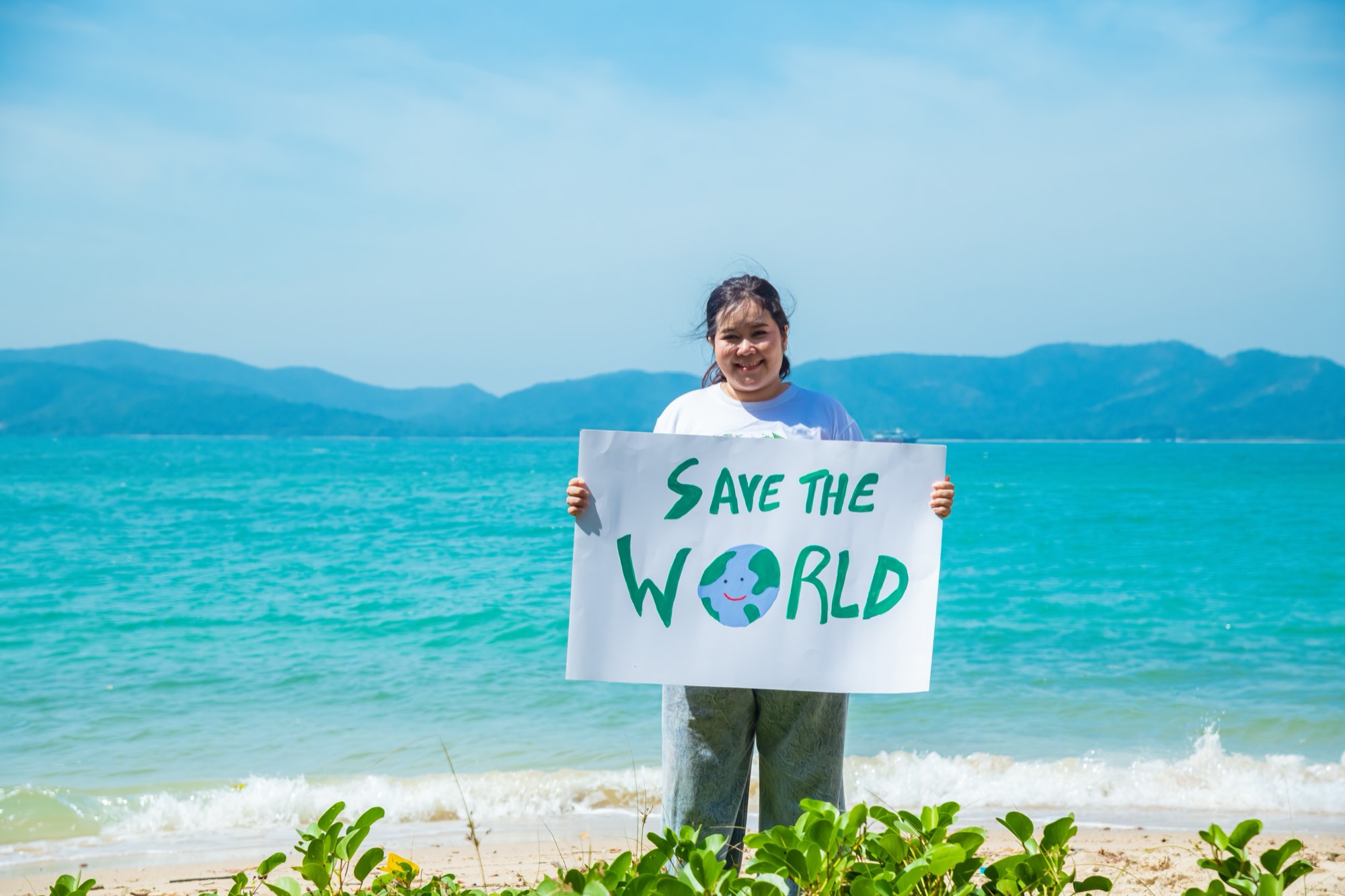 A person on a beach holding a hand-painted 'Save the World' sign